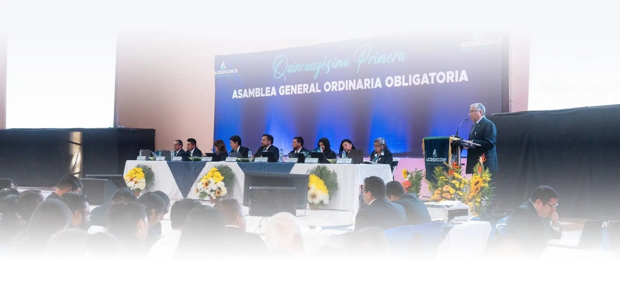 Evento formal con nueve personas sentadas en una mesa larga con micrófonos, un arreglo floral en el centro y un cartel que dice 'ASAMBLEA GENERAL ORDINARIA OBLIGATORIA'. Una persona de pie en un podio a la derecha se dirige a la audiencia.