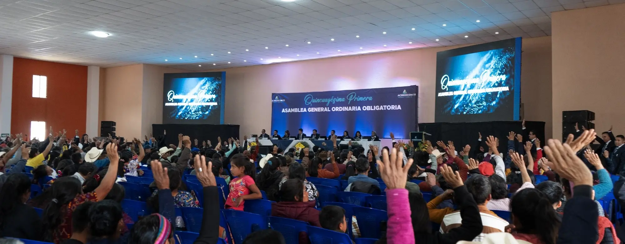 Una gran asamblea en un salón, con personas levantando la mano para participar. En el fondo, hay un escenario con una mesa larga y personas sentadas, con pantallas grandes a los lados mostrando el texto 'Asamblea General Ordinaria'.